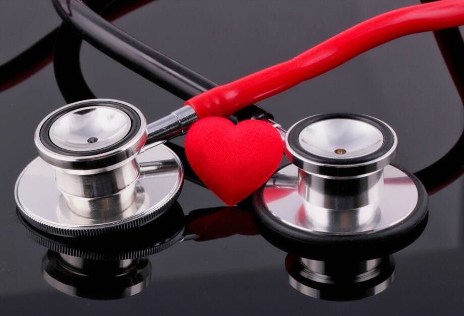Two stethoscopes and a red heart shape are placed together on a reflective black surface.