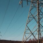 A close-up of an electricity transmission tower with power lines against a clear blue sky and distant trees.