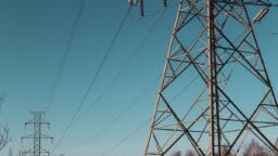 A close-up of an electricity transmission tower with power lines against a clear blue sky and distant trees.
