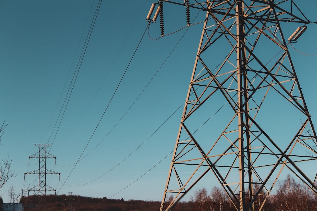 A close-up of an electricity transmission tower with power lines against a clear blue sky and distant trees.