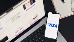 A smartphone displaying the Visa logo rests on a laptop with an online shopping website open.
