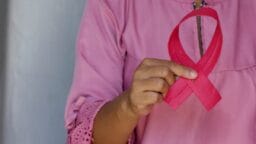 Person wearing a pink shirt holds a pink ribbon, symbolizing breast cancer awareness, against a neutral background.
