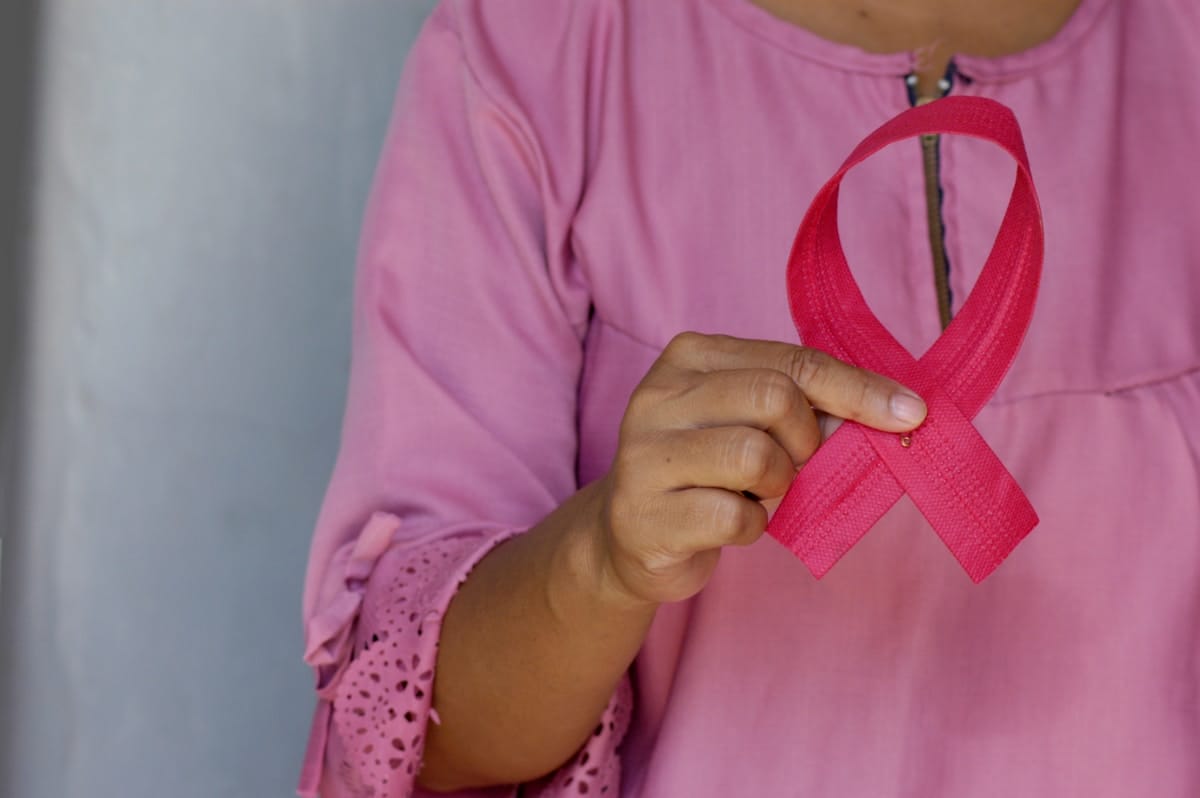 Person wearing a pink shirt holds a pink ribbon, symbolizing breast cancer awareness, against a neutral background.