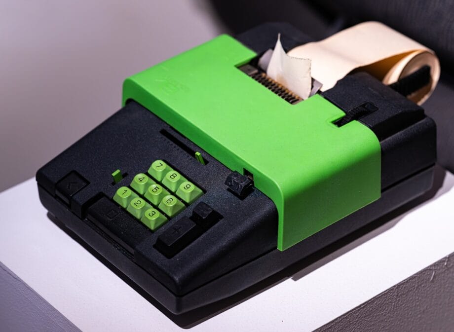 A vintage black and green calculator with a numeric keypad and a paper roll for printing calculations.