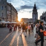 People walk through a busy European city square at sunset, with historic buildings and a clock tower in the background.