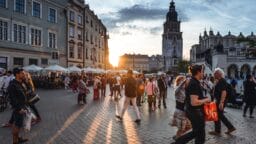 People walk through a busy European city square at sunset, with historic buildings and a clock tower in the background.