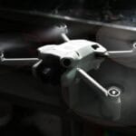 A small white quadcopter drone with spinning propellers is flying indoors near a reflective metal surface.