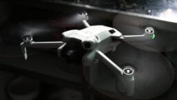 A small white quadcopter drone with spinning propellers is flying indoors near a reflective metal surface.