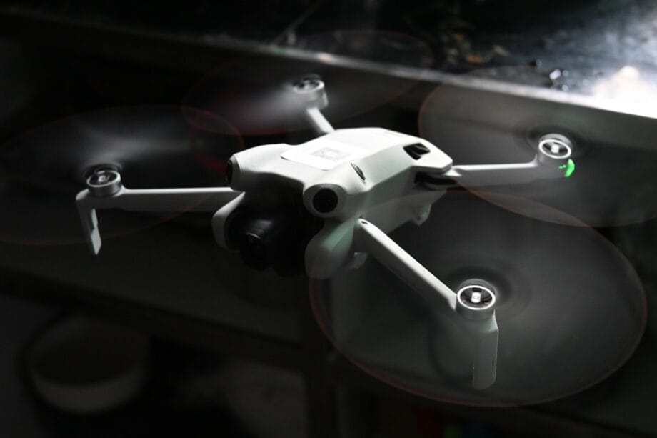 A small white quadcopter drone with spinning propellers is flying indoors near a reflective metal surface.