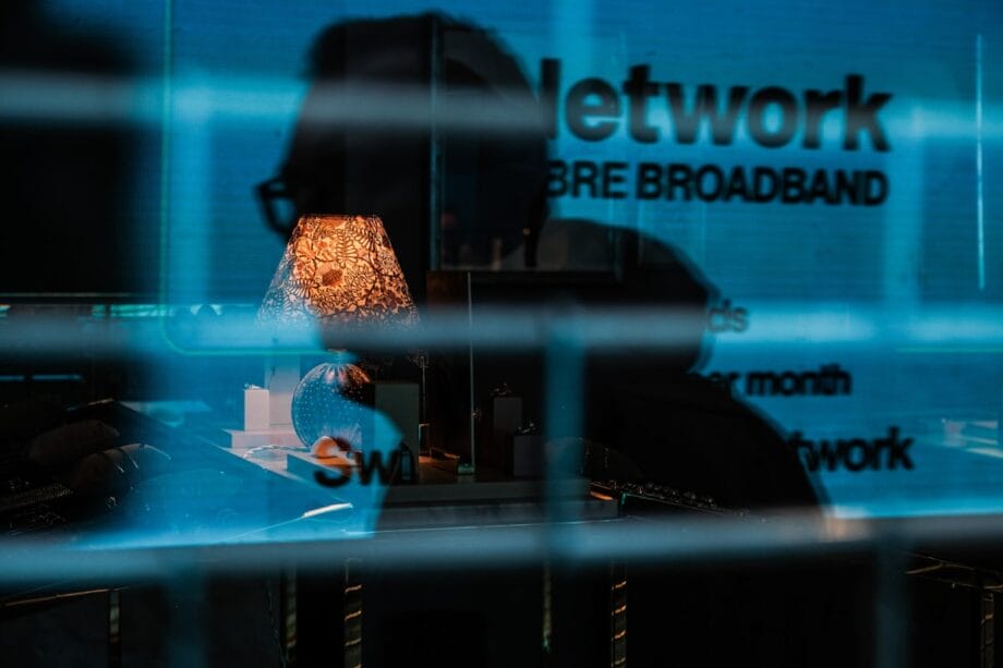 A dim lamp, desk, and various objects are seen through glass with a reflection of text reading Network and broadband.