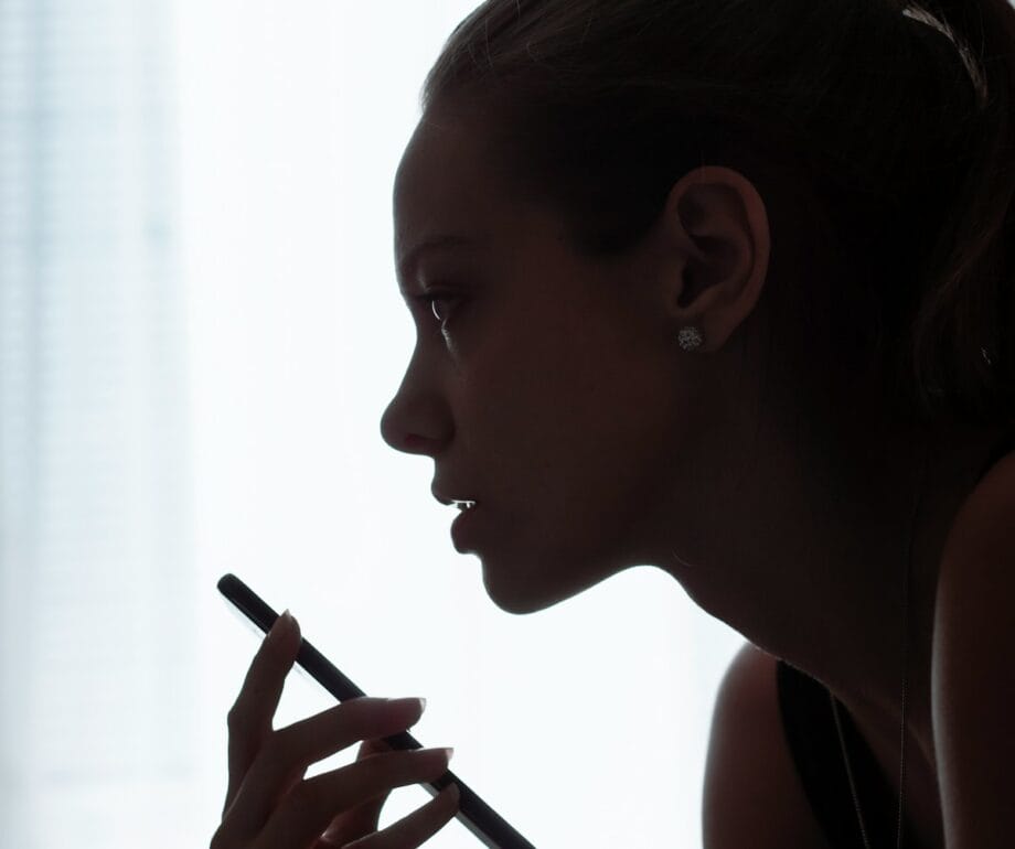 Silhouette of a woman holding a smartphone near her face, appearing to speak into it, against a bright background.