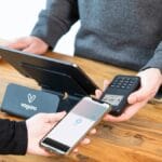 A person pays with a smartphone using contactless payment at a point-of-sale terminal on a wooden counter.