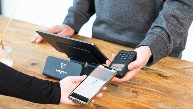 A person pays with a smartphone using contactless payment at a point-of-sale terminal on a wooden counter.
