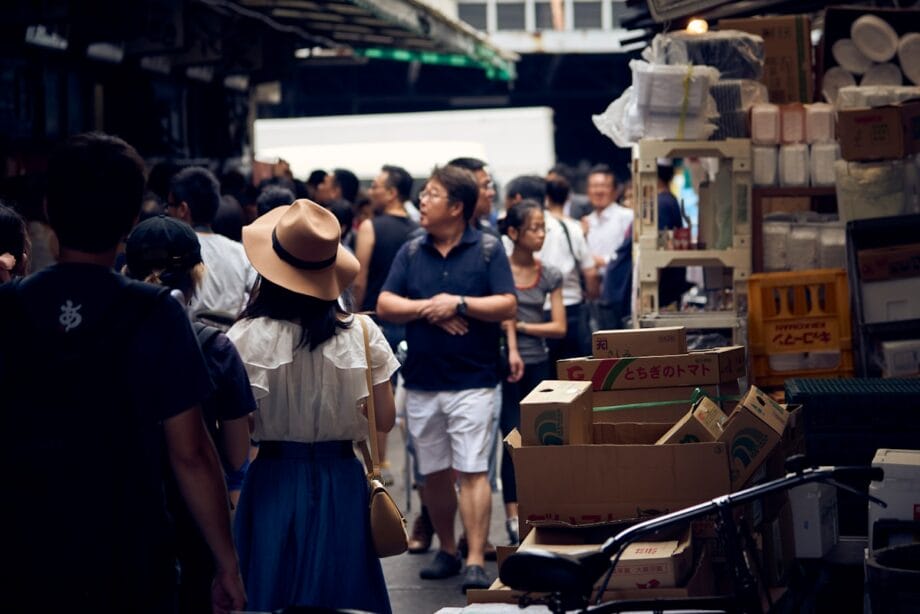 A crowded outdoor market with people walking among cardboard boxes and stalls under a covered walkway.