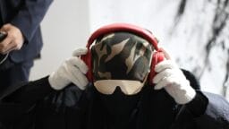 Person wearing a camouflage head covering, sunglasses, white gloves, and holding red headphones over their ears.