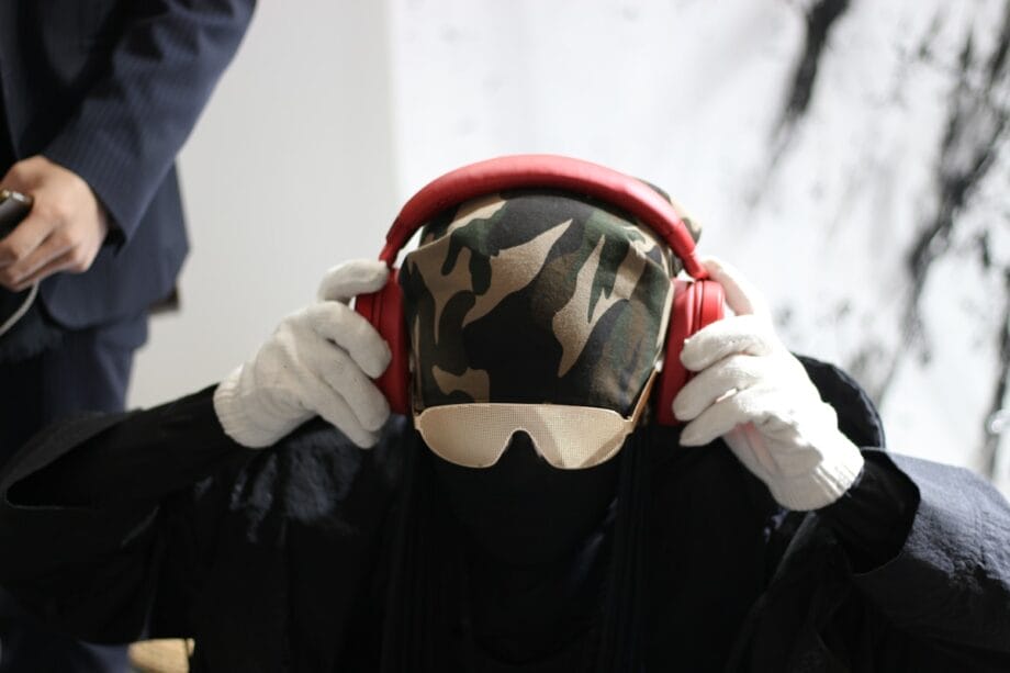 Person wearing a camouflage head covering, sunglasses, white gloves, and holding red headphones over their ears.
