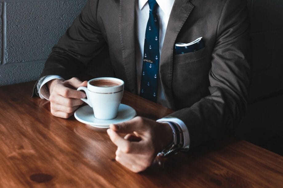 A person in a suit sits at a wooden table holding a white cup and saucer with a dark drink, possibly coffee or tea.