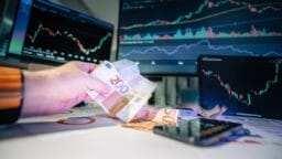 Hand counting euro banknotes on a desk with stock market charts displayed on computer screens in the background.
