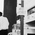 A man stands near a post with a sign that reads “NO JOBS” in an urban setting.