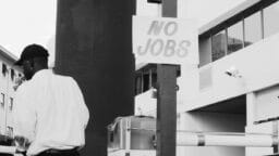 A man stands near a post with a sign that reads “NO JOBS” in an urban setting.