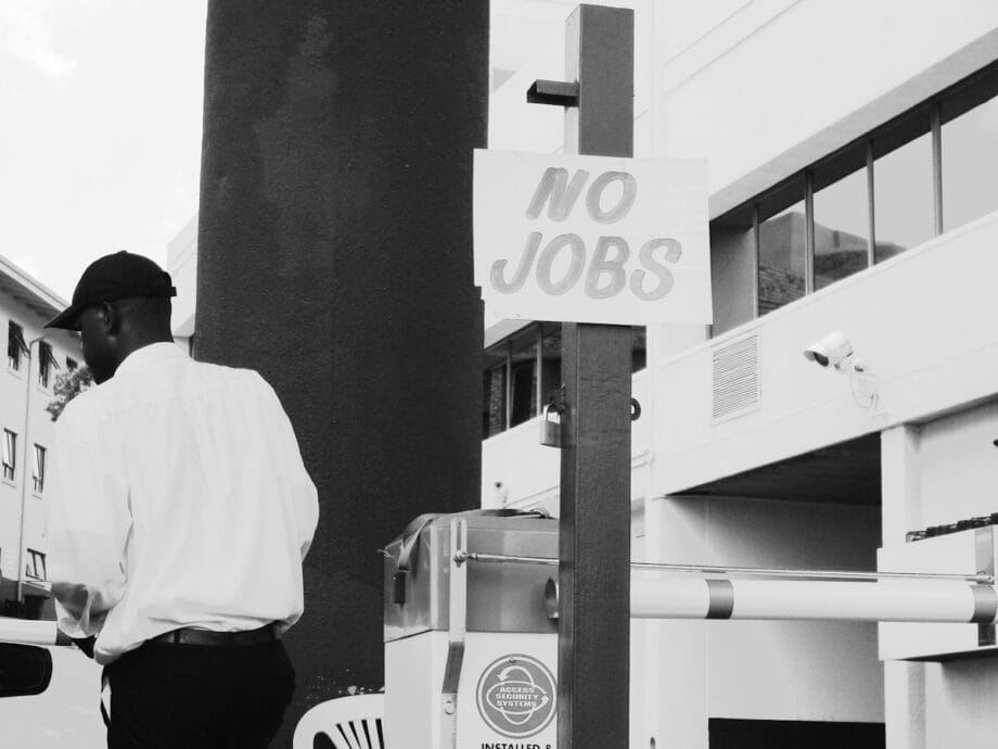 A man stands near a post with a sign that reads &ldquo;NO JOBS&rdquo; in an urban setting.