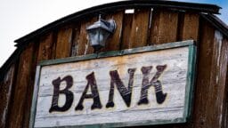 Wooden sign with the word BANK mounted on a rustic wooden building, with a lamp above the sign.