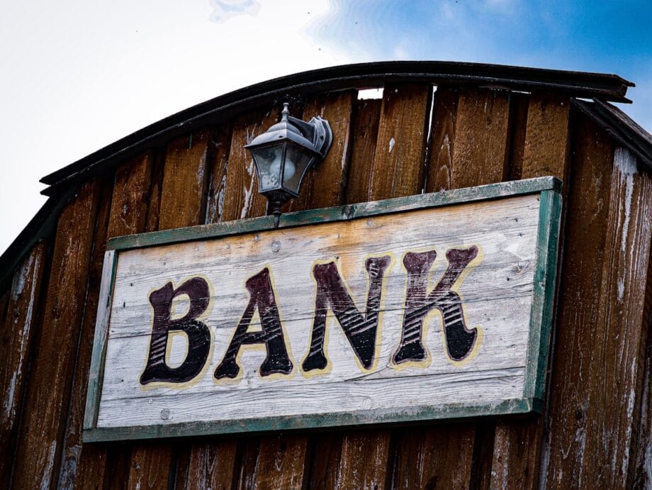 Wooden sign with the word BANK mounted on a rustic wooden building, with a lamp above the sign.