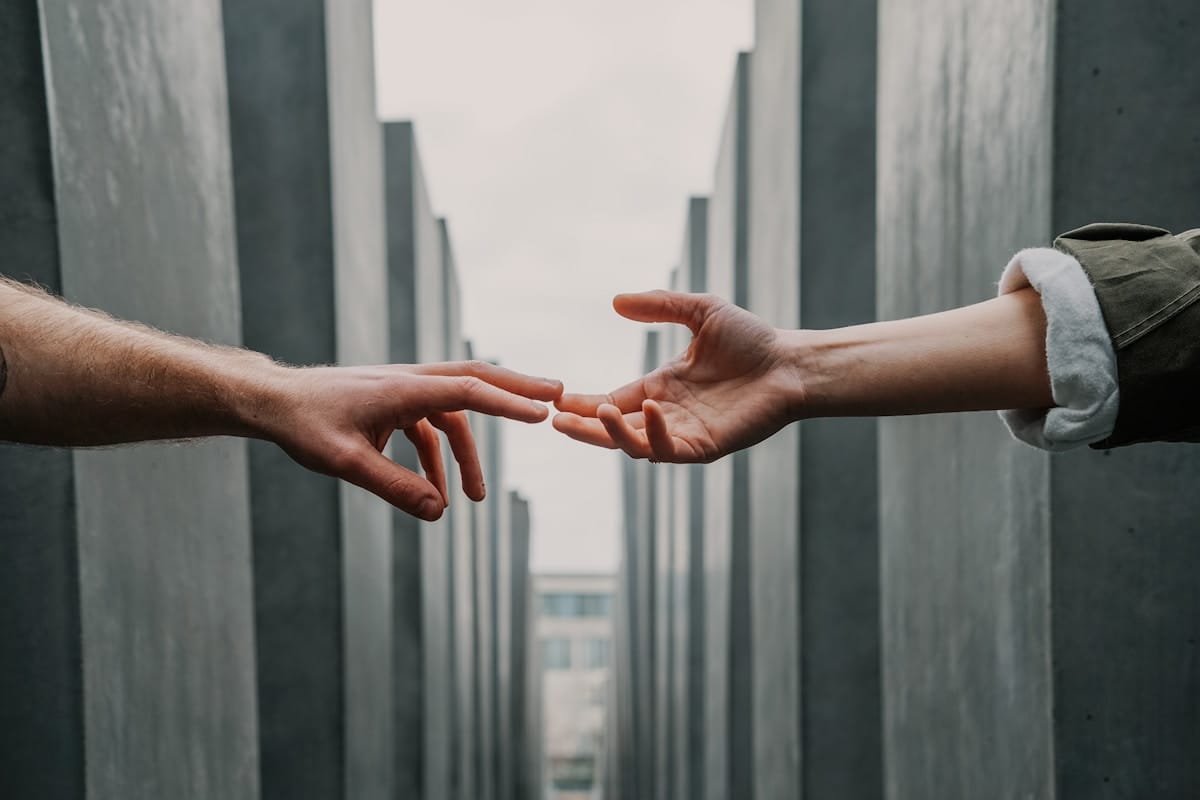 Two hands reaching toward each other in front of tall, gray concrete structures under a cloudy sky.