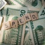 Scrabble tiles spelling SPEND rest on top of several U.S. twenty- and one-dollar bills spread out underneath.