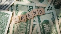 Scrabble tiles spelling SPEND rest on top of several U.S. twenty- and one-dollar bills spread out underneath.