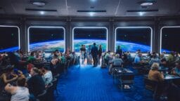 A futuristic restaurant with large windows showing a view of Earth from space, filled with diners seated at tables.