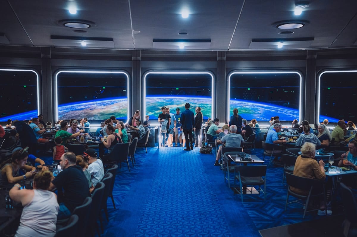 A futuristic restaurant with large windows showing a view of Earth from space, filled with diners seated at tables.