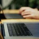 Close-up of a laptop keyboard on a wooden table with a person typing on another laptop in the blurred background.
