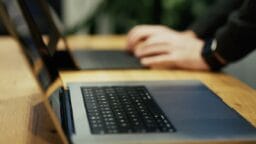 Close-up of a laptop keyboard on a wooden table with a person typing on another laptop in the blurred background.