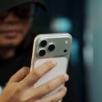 A person wearing glasses and a cap holds and looks at a smartphone with three rear cameras.