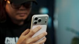 A person wearing glasses and a cap holds and looks at a smartphone with three rear cameras.