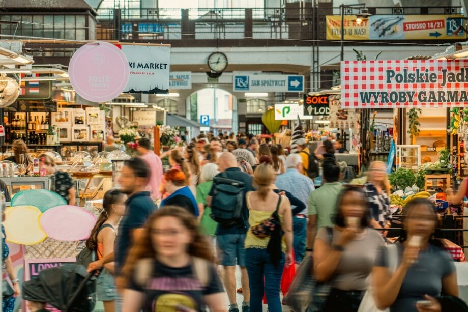 A busy indoor market filled with people walking between various food and product stalls under bright lighting.