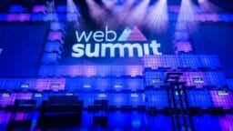 Stage setup for Web Summit conference with logo projected on a lit backdrop, empty chairs, and a podium under purple and blue lights.