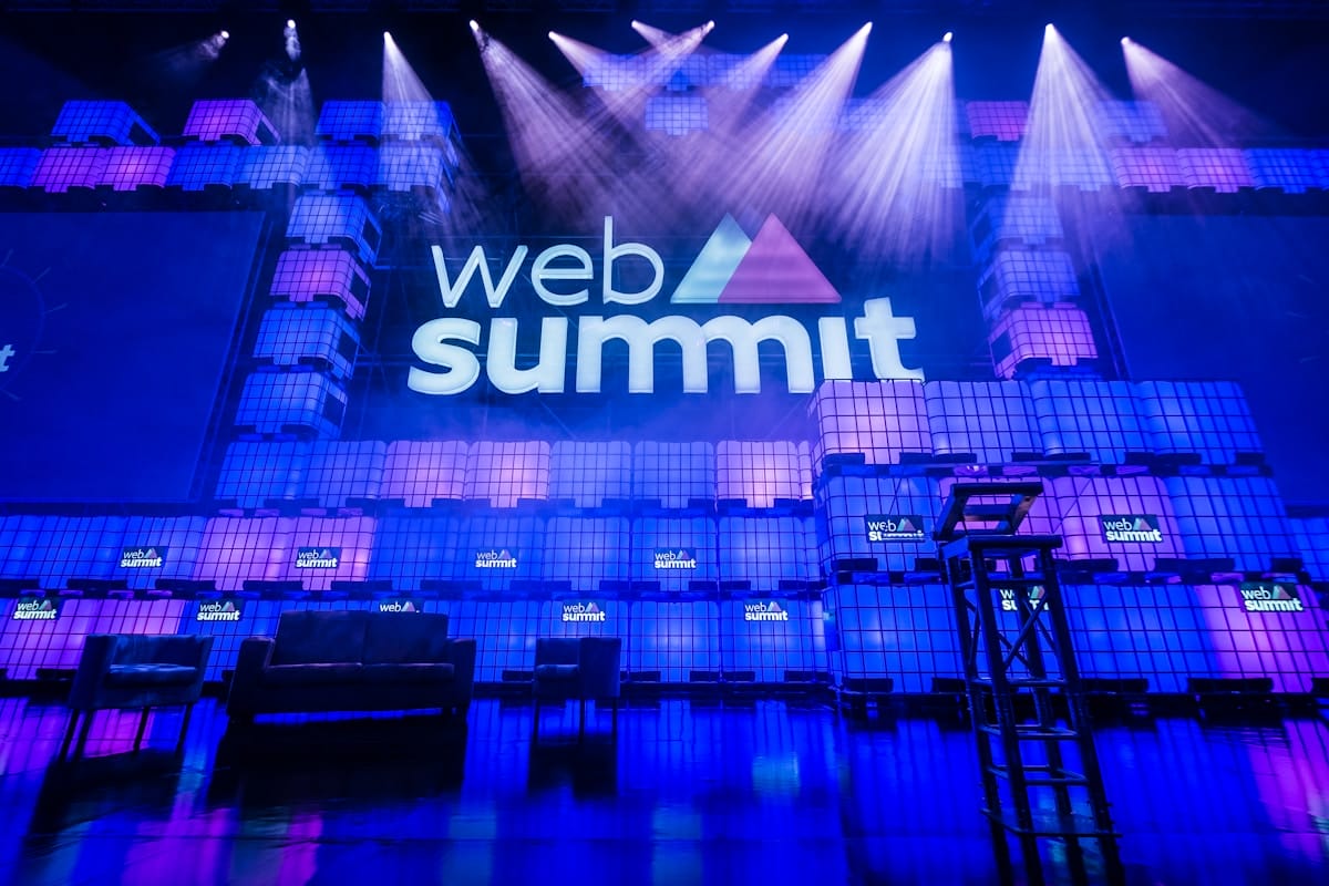Stage setup for Web Summit conference with logo projected on a lit backdrop, empty chairs, and a podium under purple and blue lights.