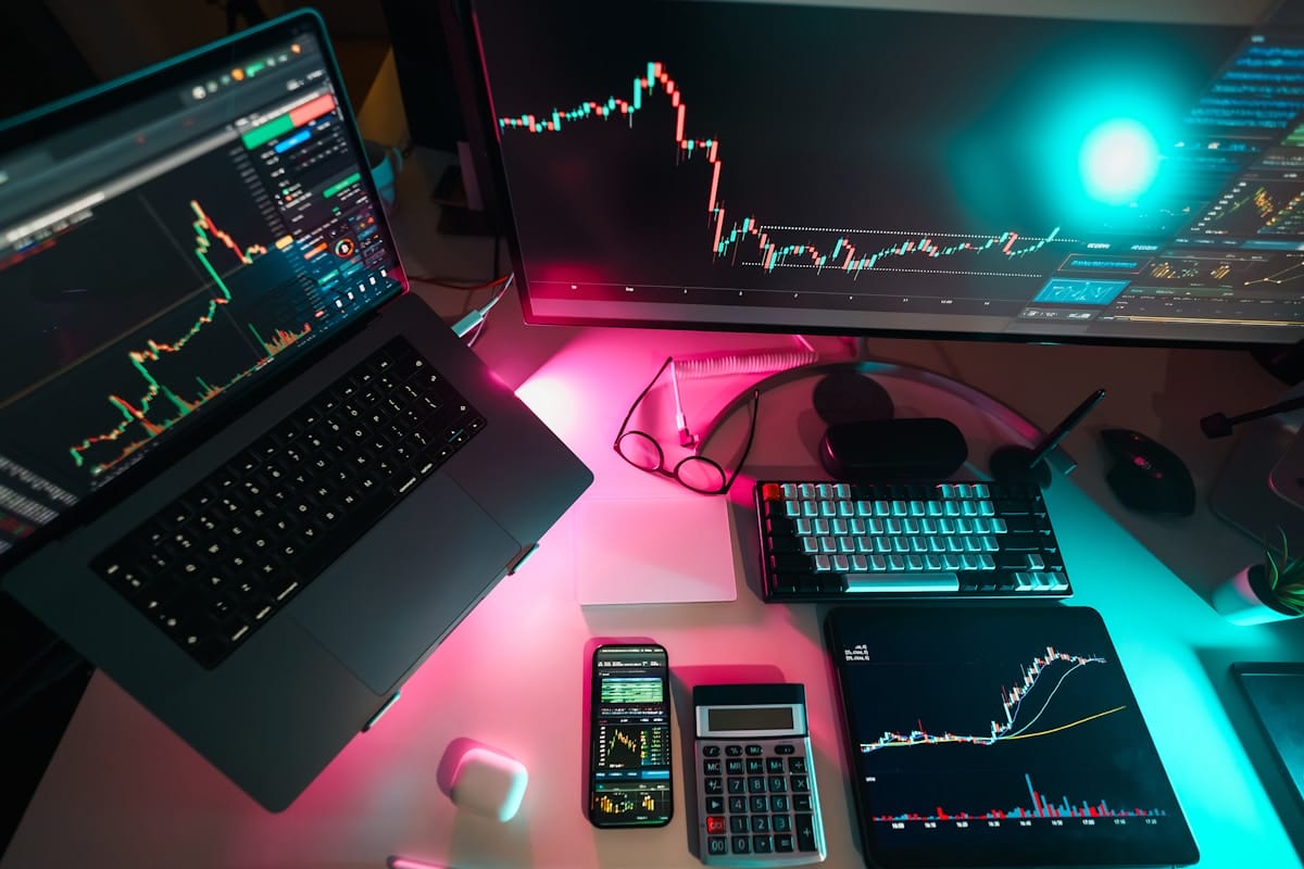 Trading workspace with charts on monitors, a laptop, smartphone, calculator, and glasses on a desk under colorful lighting.