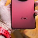 A hand holds a red Vivo smartphone with a large circular rear camera module and a wooden background.