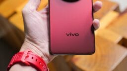 A hand holds a red Vivo smartphone with a large circular rear camera module and a wooden background.