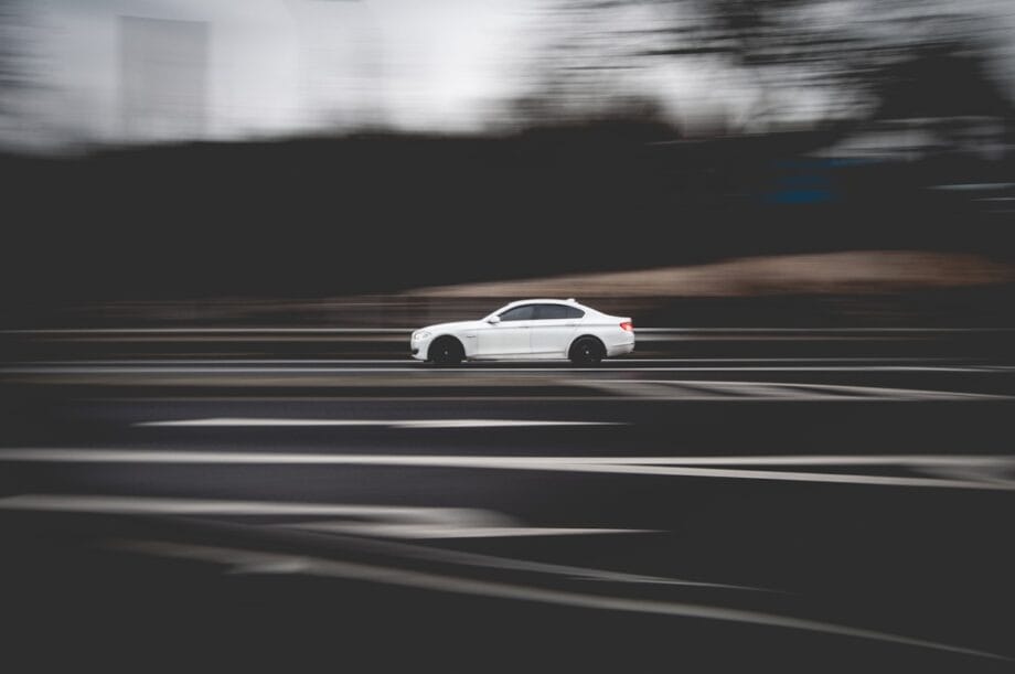 A white car is driving quickly on a multilane road, with a blurred background indicating motion.