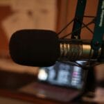 Close-up of a studio microphone suspended on a boom arm, with a blurred background of papers and a laptop.
