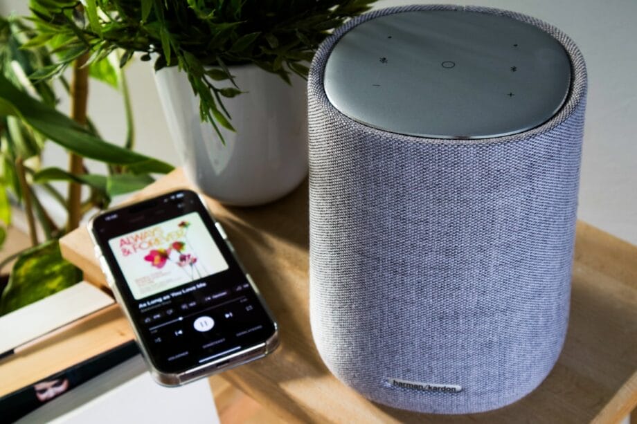A Harman Kardon smart speaker sits on a wooden surface next to a smartphone displaying a music app.