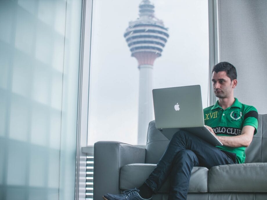 Man sitting on a couch using a laptop with a tall tower visible through the window in the background.