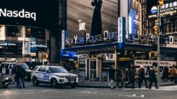 Pedestrians cross a street near NYPD vehicles and a New York Police Department station in a busy city area with electronic billboards.