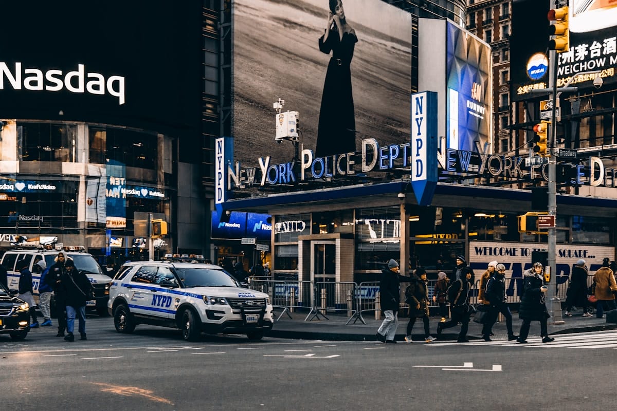 Pedestrians cross a street near NYPD vehicles and a New York Police Department station in a busy city area with electronic billboards.