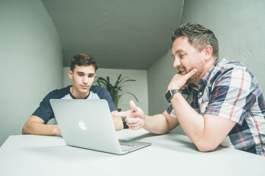 Two men sitting at a table looking at a laptop screen, one pointing at the screen while the other listens.