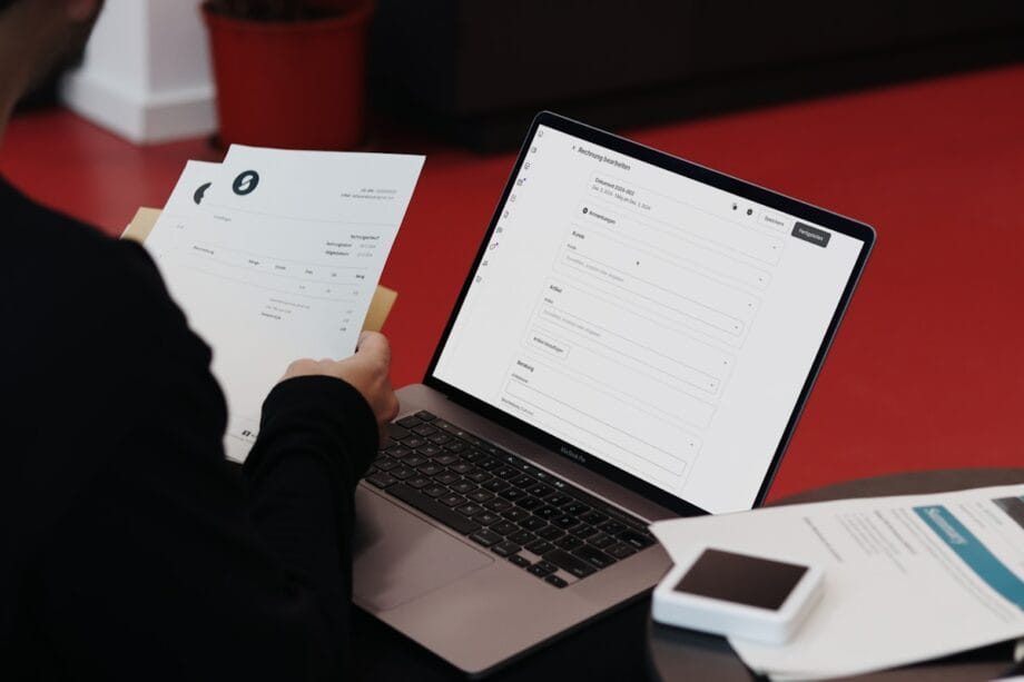 Person reviewing a printed document while filling out an online form on a laptop at a desk with papers and a smartphone.
